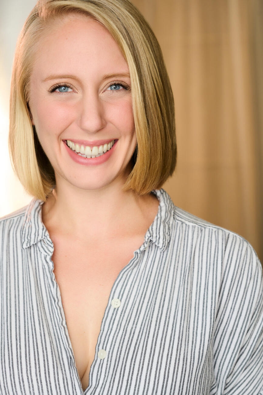 Nicole smiles broadly at the camera. She is a white woman with blue eyes and a blonde bob. She wears a white and gray striped shirt.